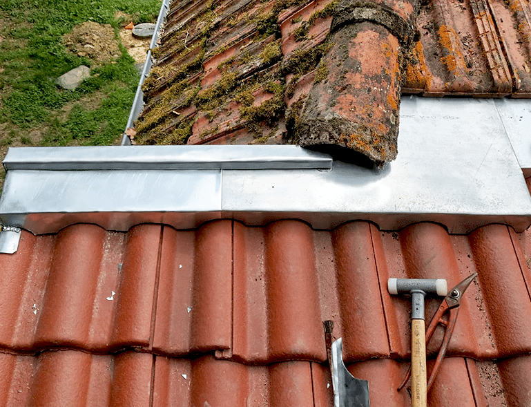 Toiture en réparation avec tuiles anciennes et neuves, solin métallique et outils posés sur les tuiles.