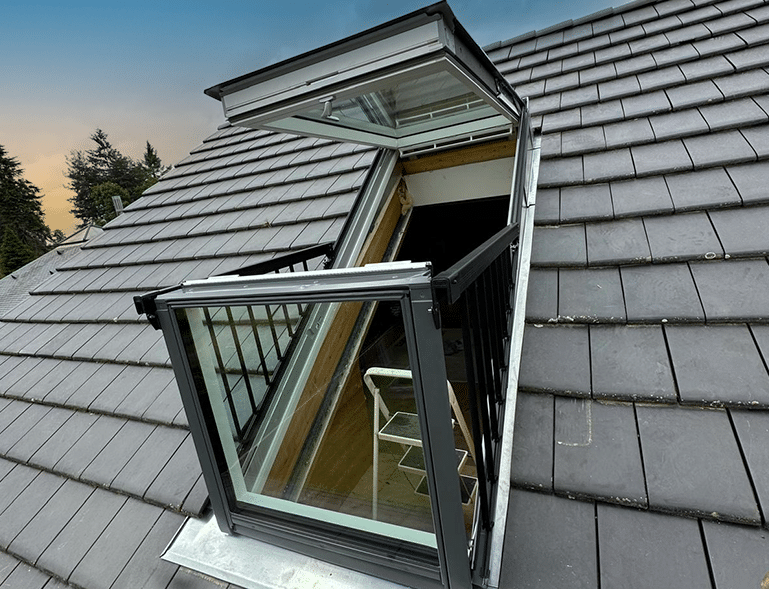 fenêtre de toit Velux cabrio ouverte en position balcon sur toiture en tuile béton anthracite au coucher de soleil