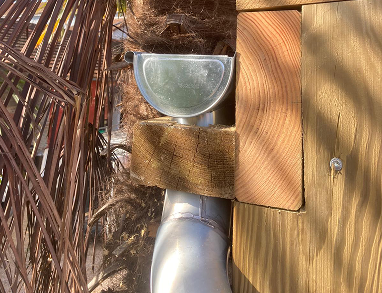 Tuile et gouttière en métal fixées à une façade en bois, avec des feuilles de palmier sèches à gauche.