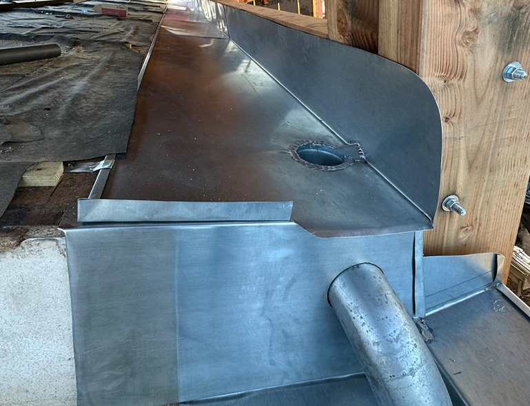 Détail d’un solin métallique nouvellement posé autour d’un poteau en bois, avec une ouverture circulaire et une conduite verticale traversant le solin; matériaux de construction visibles autour.